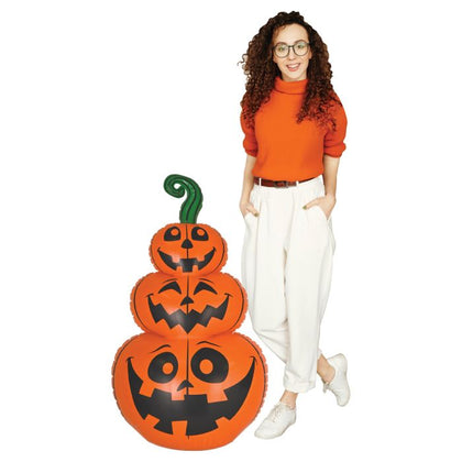 Woman in an orange sweater standing next to a large inflatable pumpkin with jack-o'-lantern faces on a white background