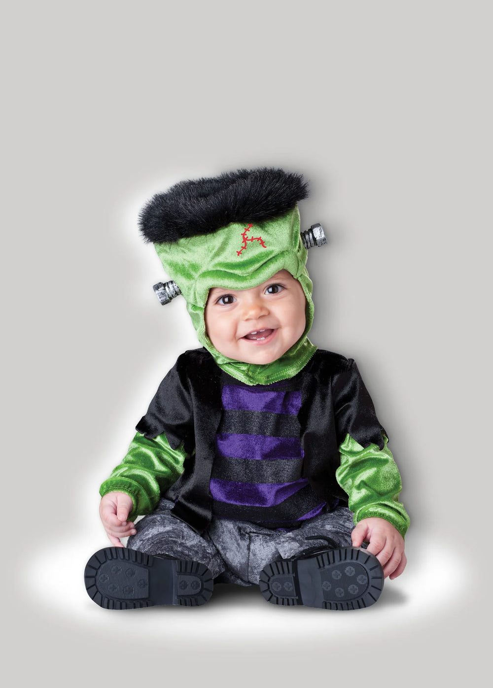 A baby wearing a Frankenstein costume, sitting against a solid gray background. The costume includes a green headpiece with black hair and silver bolts, a black and purple striped shirt, and green striped sleeves, with gray pants and black booties.