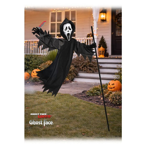 A Ghostface Halloween decoration costume shown standing on a lawn in front of a house. The figure wears a black hooded robe with a white face mask featuring black eyes and a white mouth, holds a black staff, and has one arm extended with a red knife prop.
