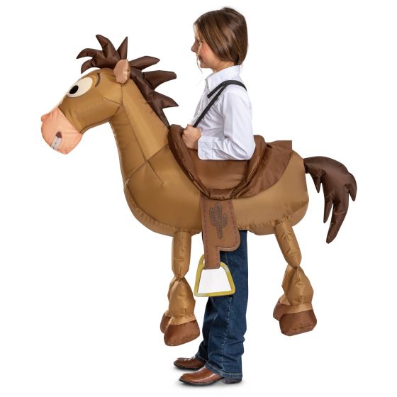A child wearing a Toy Story-style cowboy horse costume, shown from the side against a plain white background. The costume includes a tan and brown inflatable horse body with a saddle, bridle, and dangling white-and-yellow bell, and the child is dressed in a white shirt and blue jeans.