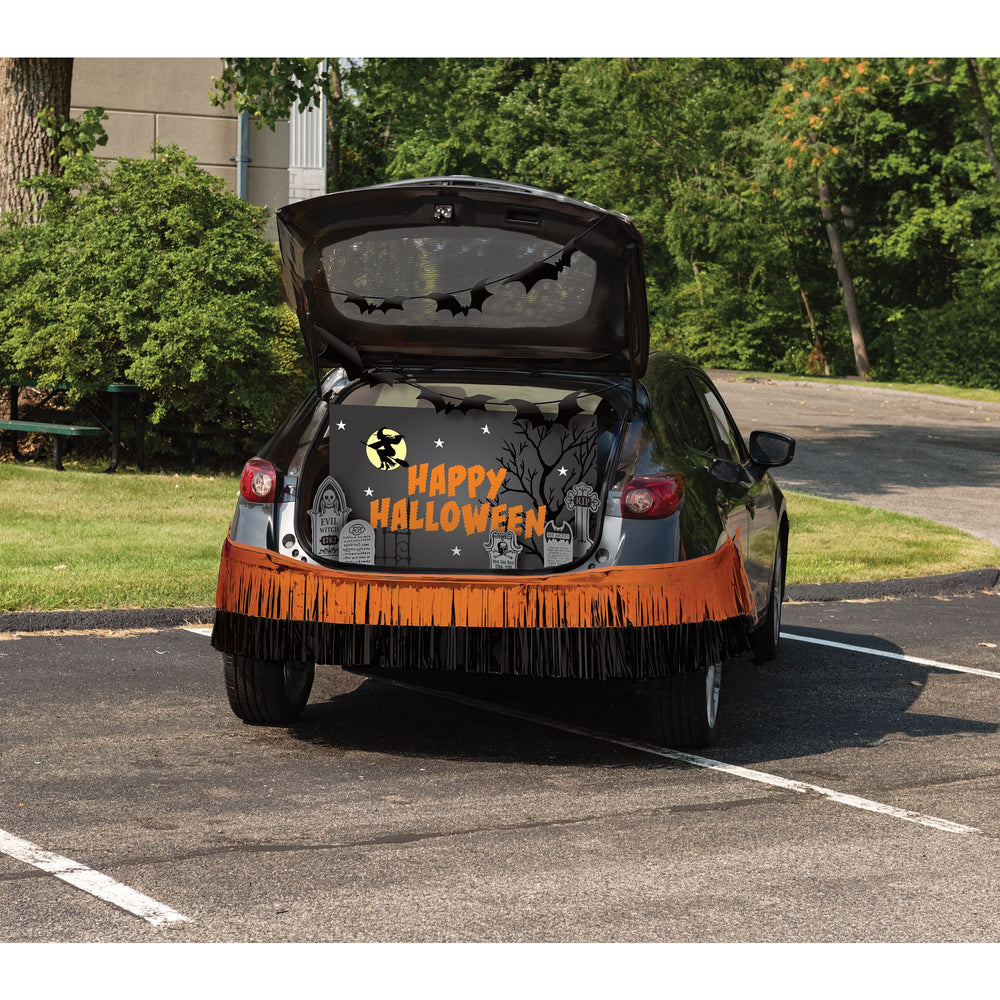 A black car parked in a lot with its trunk open, decorated for Halloween. The back of the car features a black Halloween-themed fabric cover with a fringe bottom, and the words 'HAPPY HALLOWEEN' in orange and white.