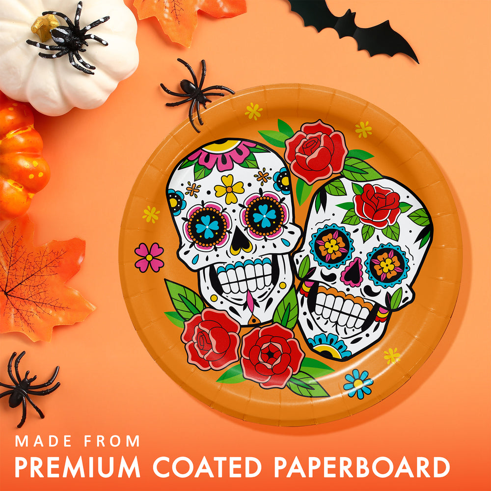 A round orange paper plate with a colorful Day of the Dead sugar skull and floral design, shown on an orange background with Halloween props including pumpkins, leaves, and black spiders. Text at the bottom reads 'MADE FROM PREMIUM COATED PAPERBOARD'.