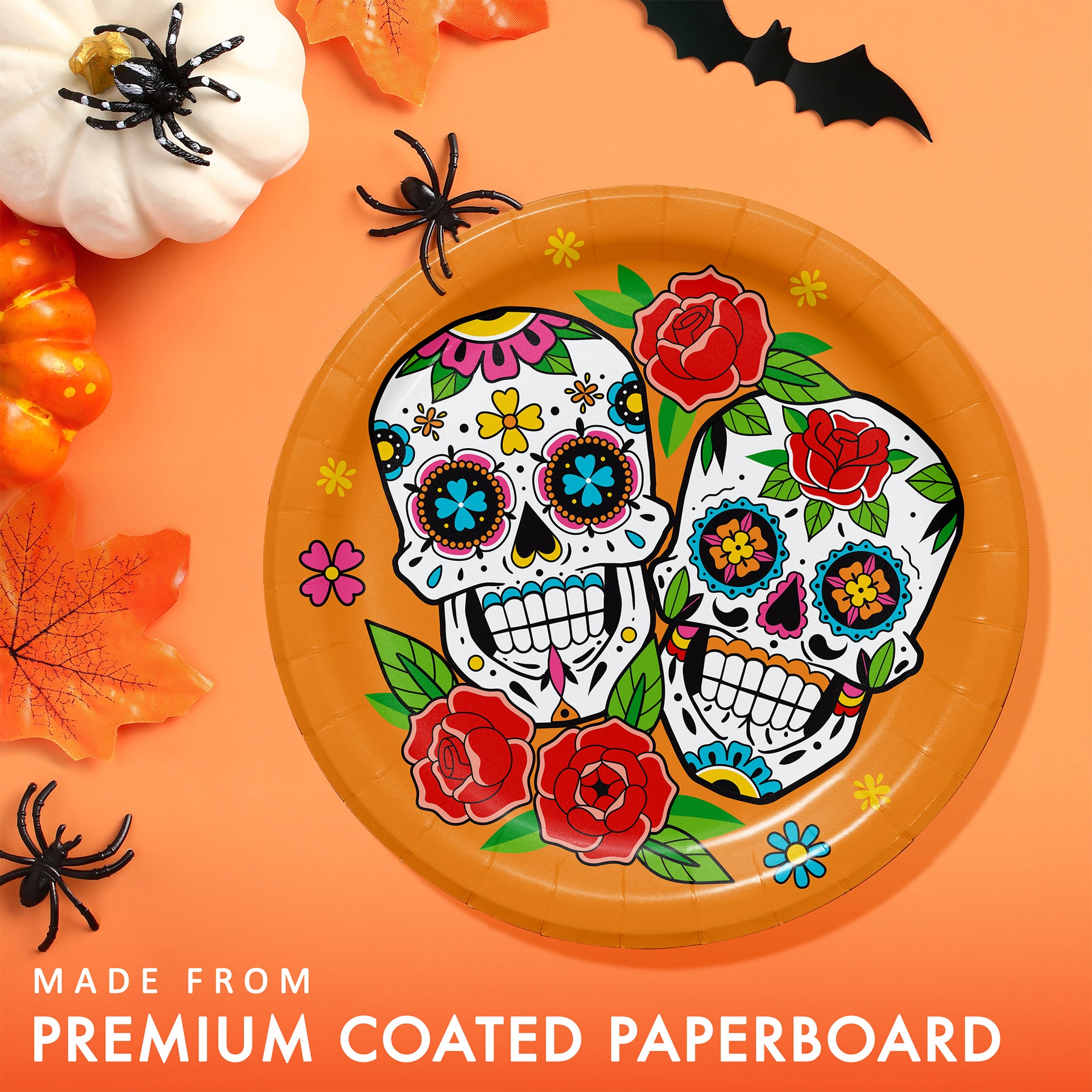 A round orange paper plate with a colorful Day of the Dead sugar skull and floral design, shown on an orange background with Halloween props including pumpkins, leaves, and black spiders. Text at the bottom reads 'MADE FROM PREMIUM COATED PAPERBOARD'.