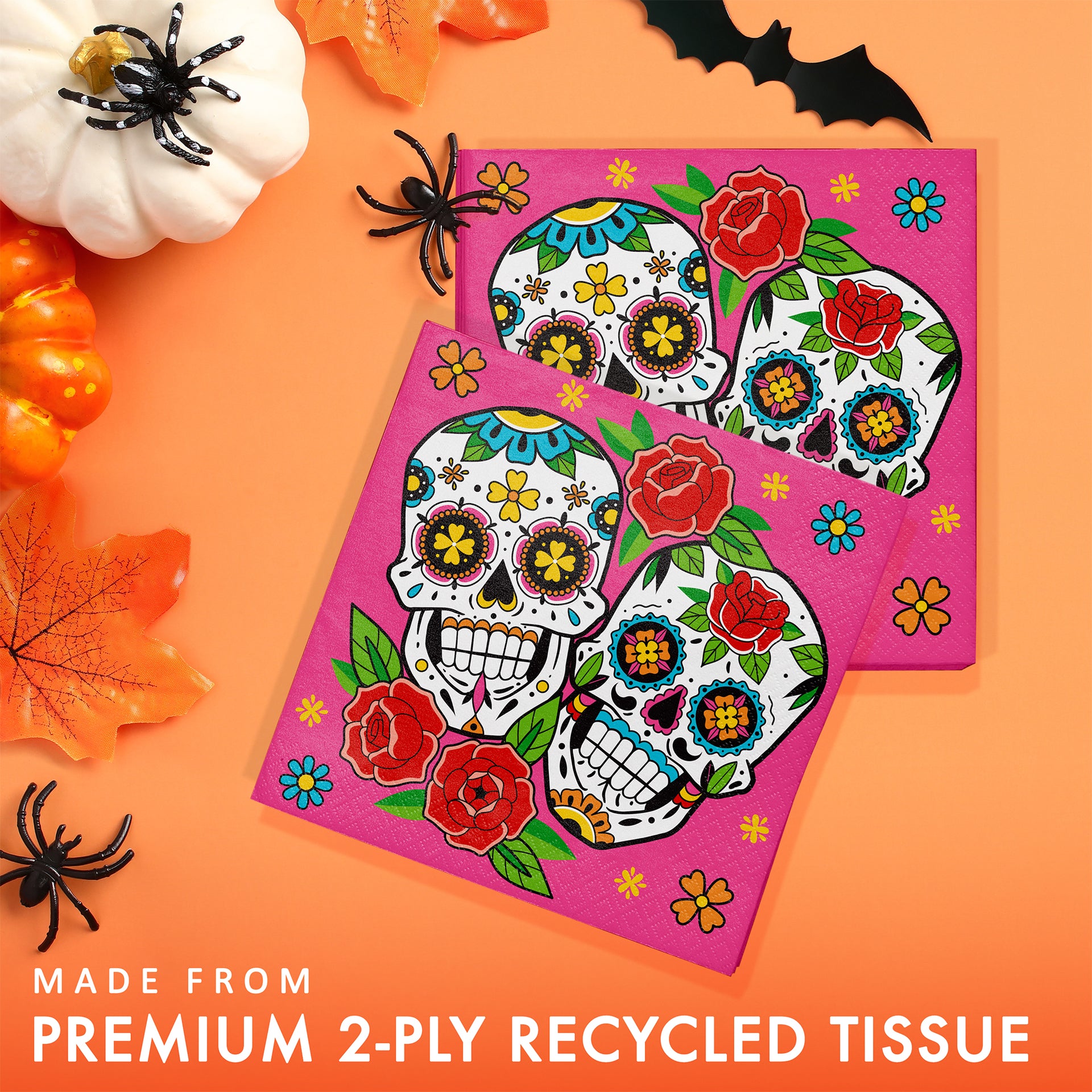 Two square pink tissue napkins with colorful sugar skull and floral designs, arranged on an orange background with Halloween props and text reading 'MADE FROM PREMIUM 2-PLY RECYCLED TISSUE'.