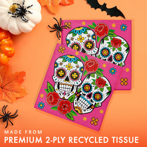 Two square pink tissue napkins with colorful sugar skull and floral designs, arranged on an orange background with Halloween props and text reading 'MADE FROM PREMIUM 2-PLY RECYCLED TISSUE'.