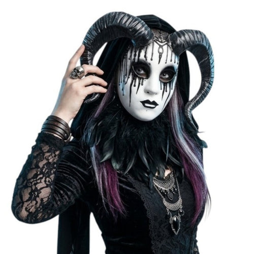 A person wearing a black and white gothic mask with long curved black horns, black lace clothing, layered silver necklaces, and dark purple-streaked hair, posed against a plain white background.