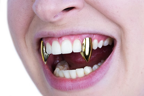 Close-up of a person's mouth wearing two gold vampire fang teeth guards over white teeth, set against a plain white background.