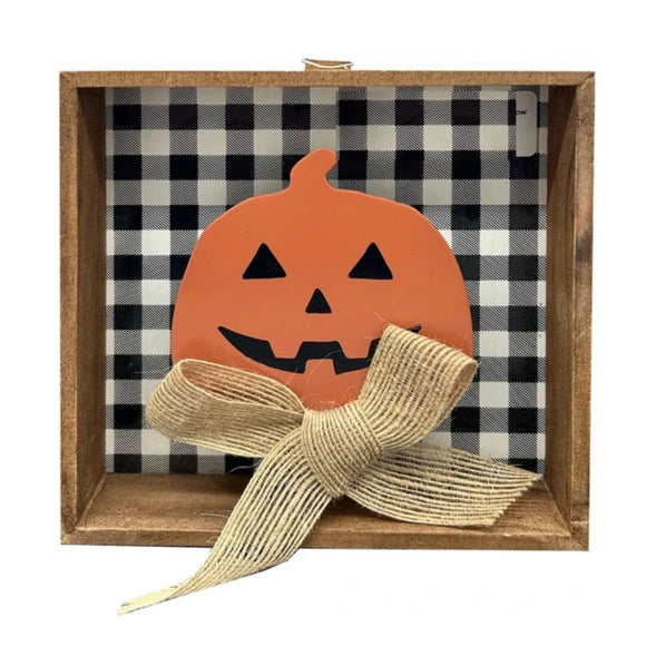 A square wooden shadow box decoration featuring a black and white buffalo plaid fabric lining, an orange jack-o'-lantern cutout with black facial features, and a large beige burlap ribbon bow tied in front.