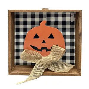 A square wooden shadow box decoration featuring a black and white buffalo plaid fabric lining, an orange jack-o'-lantern cutout with black facial features, and a large beige burlap ribbon bow tied in front.