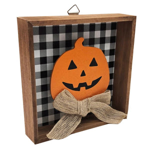 A square wooden shadow box wall decor piece featuring a black and white buffalo plaid background, an orange felt jack-o'-lantern face, and a large beige burlap ribbon bow at the front.