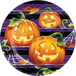 A round Halloween-themed plate or platter featuring three carved orange pumpkins with green stems and leaves, set against a purple brick wall background with black stripes, white cobwebs, and scattered autumn leaves.