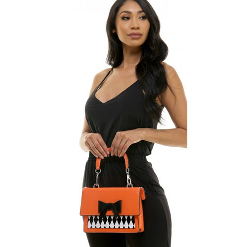 A woman wearing a black jumpsuit holds a bright orange handbag with a black bow and a black-and-white diamond pattern on the front, set against a plain white background.