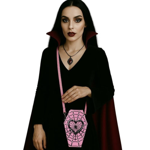 A woman with long black hair and gothic makeup wears a black vampire-style costume dress with a dark red cape. She is holding a pink coffin-shaped crossbody bag with a black spiderweb pattern and a black heart in the center.