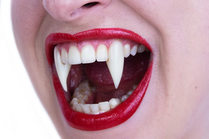 Close-up of a person's mouth wearing a vampire fang tooth set, with bright red lipstick and white pointed fake teeth visible against a plain white background.