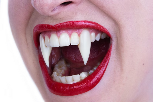 Close-up of a person's mouth wearing a vampire fang tooth set, with bright red lipstick and white pointed fake teeth visible against a plain white background.