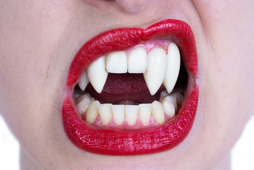 A close-up of a person's mouth with bright red lipstick, showing pointed fangs and a full set of natural teeth against a white background.