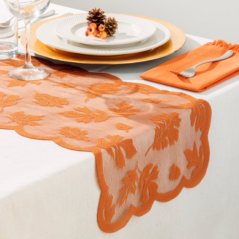 An orange lace table runner with scalloped edges draped across a white table, styled with white plates, gold charger plates, a wine glass, and an orange napkin with a silver fork.