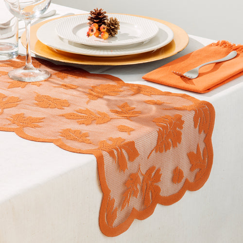 An orange lace table runner with scalloped edges draped across a white table, styled with white plates, gold charger plates, a wine glass, and an orange napkin with a silver fork.