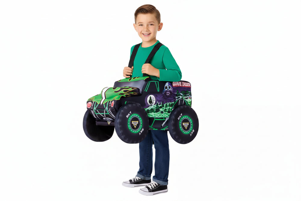 A young boy wearing a Monster Trucks 'Grave Digger' costume stands behind a toy ride-on truck, holding the handle. The costume includes a black cap with a 'Monster Trucks' logo, a green long-sleeve shirt, and blue jeans.