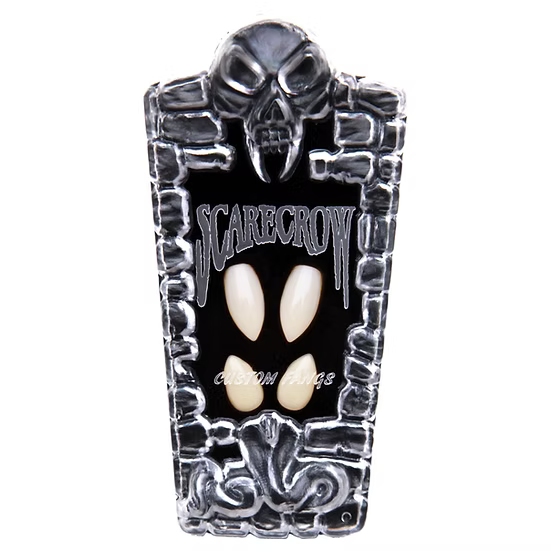 A coffin-shaped Halloween mask or plaque featuring a black background, silver skull and stone-like border details, and two off-white pointed teeth-like cutouts in the center. The word 'SCARECROW' is printed across the top.