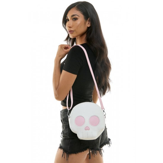 A woman wearing a black t-shirt and denim shorts carries a white skull-shaped crossbody bag with a pink strap and pink eye details against a white background.