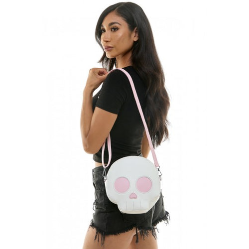A woman wearing a black t-shirt and denim shorts carries a white skull-shaped crossbody bag with a pink strap and pink eye details against a white background.