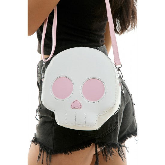 A white faux-leather skull-shaped crossbody bag with pink eye sockets and nose, worn by a model in a black top and denim shorts.