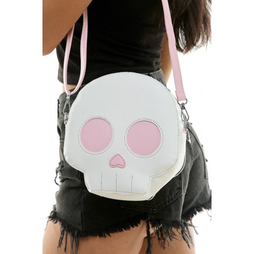 A white faux-leather skull-shaped crossbody bag with pink eye sockets and nose, worn by a model in a black top and denim shorts.