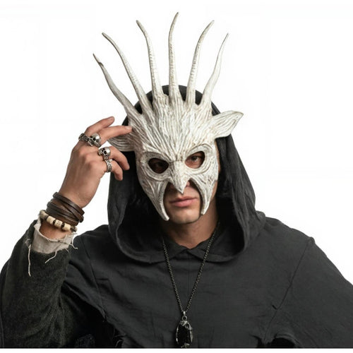 A person wearing a detailed white horned fantasy mask and a black hooded robe, with rings and bracelets on one hand, against a plain white background.