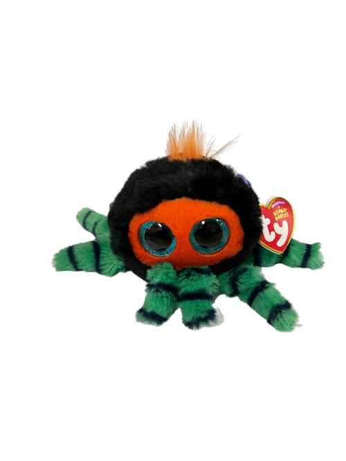 A Ty Beasty plush spider toy with a black body, orange face, green striped legs, and orange hair, resting on white papers on a wooden desk with a hand holding one corner.