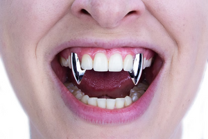 A close-up of a person's mouth wearing two shiny silver vampire fangs on the upper front teeth.