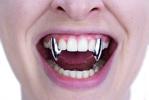 A close-up of a person's mouth wearing two shiny silver vampire fangs on the upper front teeth.