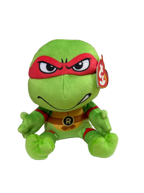 A green Teenage Mutant Ninja Turtles plush toy with a red mask and yellow chest emblem, sitting on a white sheet of paper on a wooden surface.